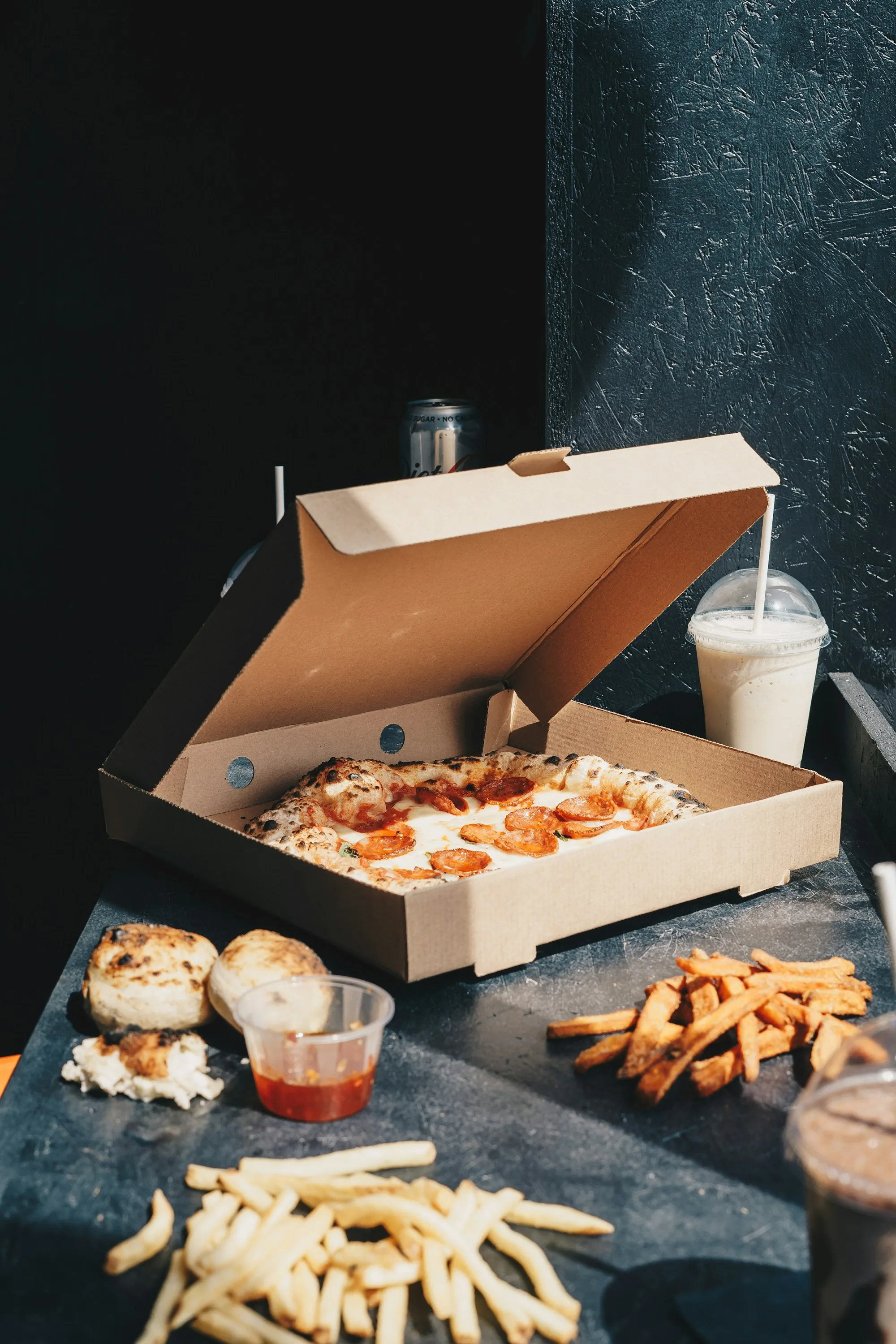 A pizza takeout box with fries, sauce, and a shake on a moody dark counter — the takeout / online order moment, packaged and ready